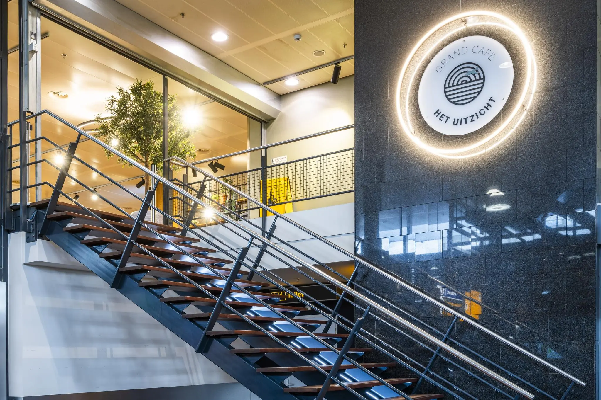 Trap naar Grand Café Het Uitzicht op Rotterdam The Hague Airport met modern interieur en verlichte gevel.